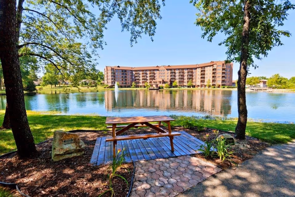 A peaceful outdoor scene at Lakeview Village featuring a wooden picnic table on a small wooden platform surrounded by trees and plants. In the background, there is a large pond with a fountain and a multi-story residential building reflecting in the water under a clear blue sky.