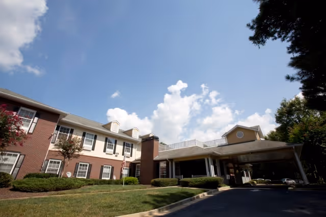 Front exterior of a two-story senior living facility with a covered porte-cochere, landscaped lawn, and blue sky.