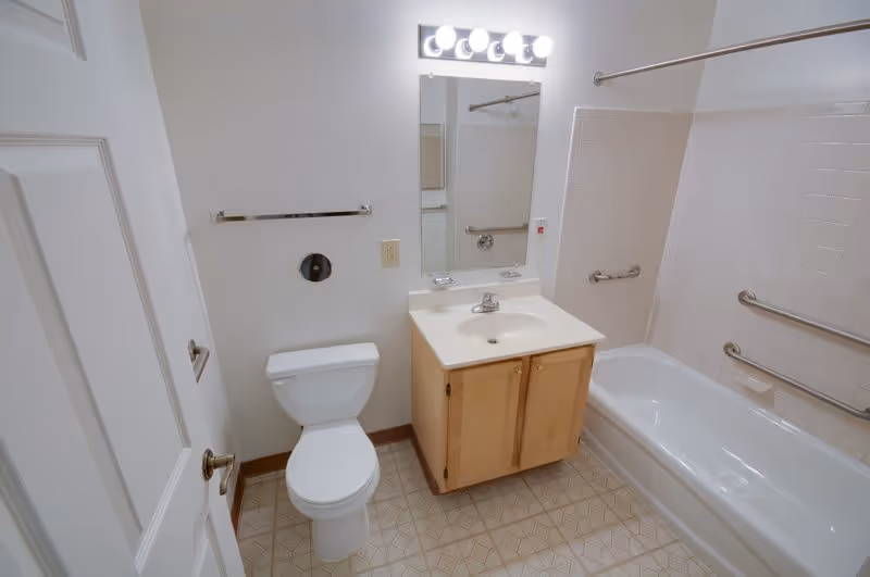 A clean bathroom with a white toilet, a wooden vanity with a sink, a mirror above the sink with four light bulbs, and a bathtub with white tiled walls and multiple grab bars installed for safety.