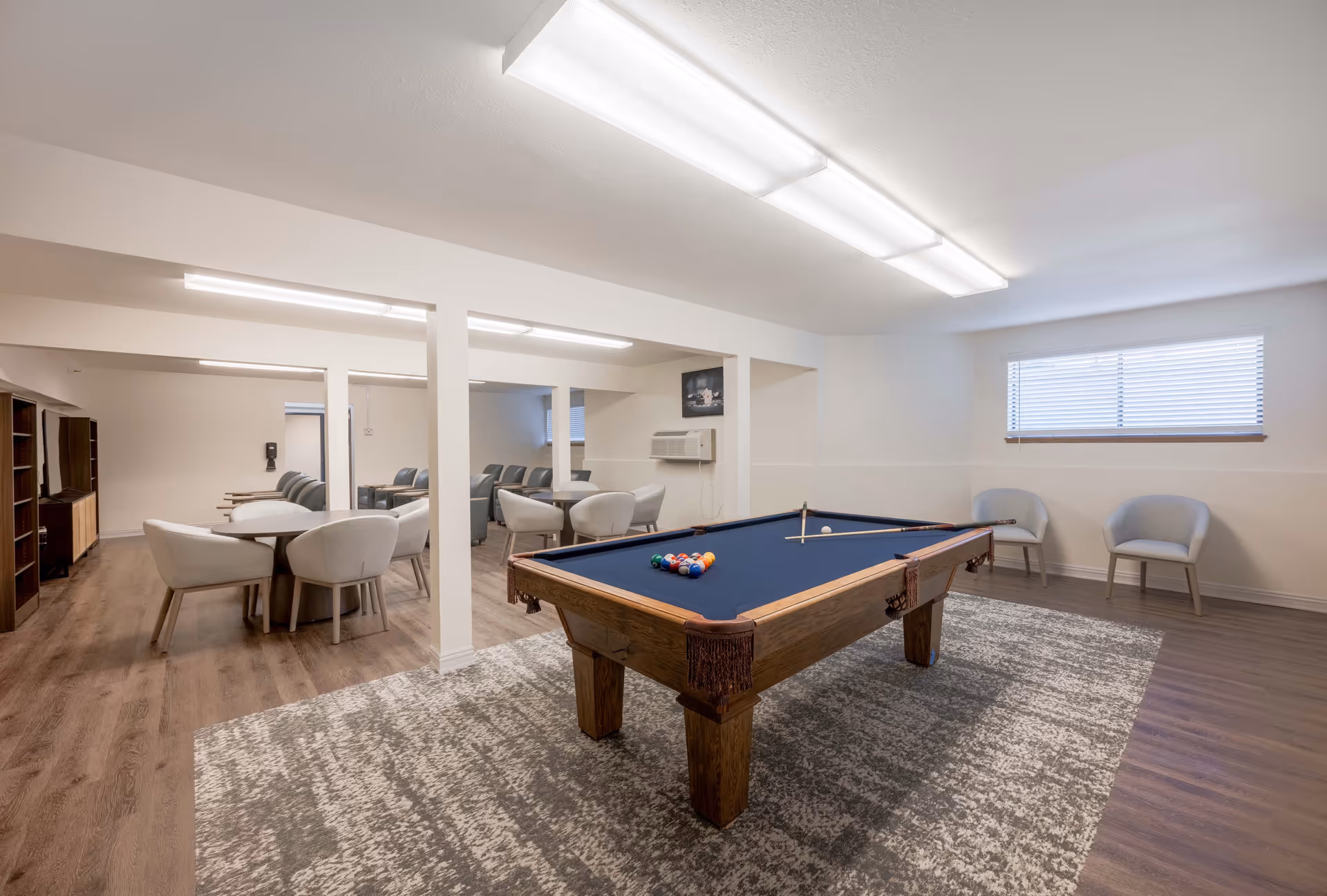 A spacious recreational room with a pool table in the center on a patterned rug. The room has light-colored walls and wood flooring. There are several seating areas with modern chairs and tables, and a window with blinds letting in natural light. The room is well-lit with ceiling lights and has an air conditioning unit mounted on the wall.
