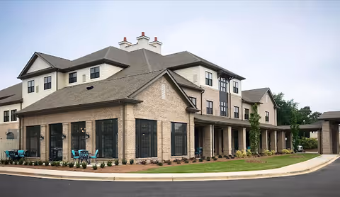 Exterior view of a multi-story senior living facility building with beige brick and siding, large windows, and a covered entrance. There are outdoor seating areas with turquoise chairs and small tables on a landscaped lawn with shrubs and a paved driveway in front.