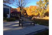 A wooden bench on a paved patio in front of a building with colorful autumn trees in the background.