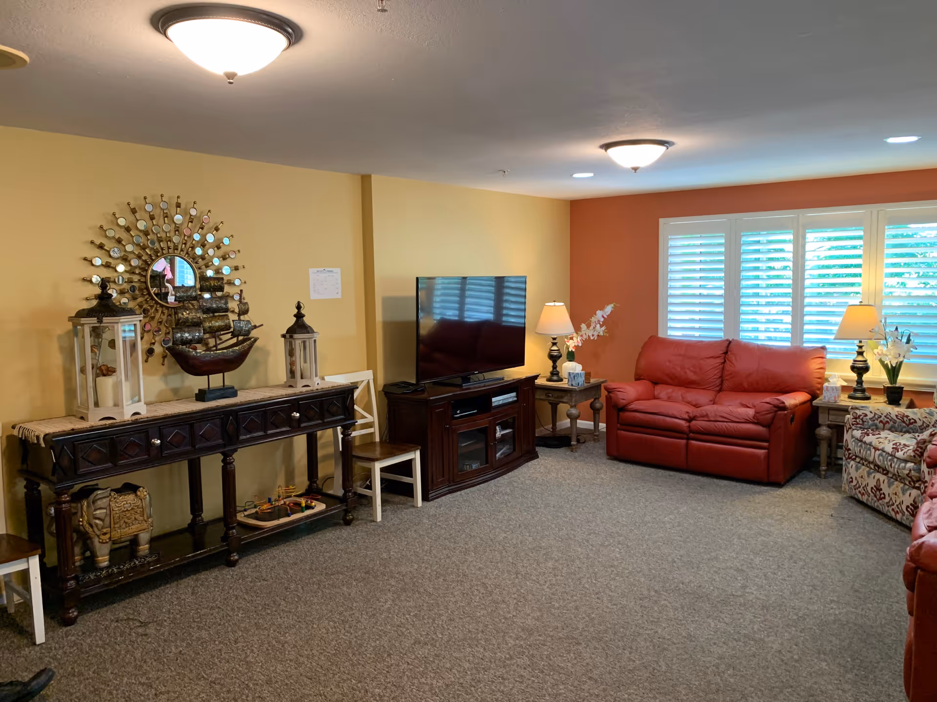 A cozy living room with beige and orange walls, featuring a red leather loveseat, a patterned armchair, a wooden TV stand with a flat-screen TV, two side tables with lamps and flowers, a decorative console table with lanterns and a ship model, and a round mirror with small circular mirrors around it. The room is carpeted and has white plantation shutters on the windows.