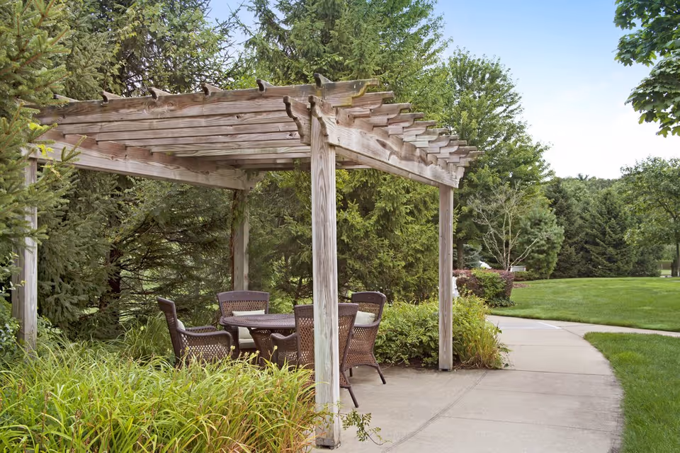 Outdoor seating area with a wooden pergola covering a round table and four chairs, surrounded by green bushes, trees, and a concrete pathway in a garden setting.