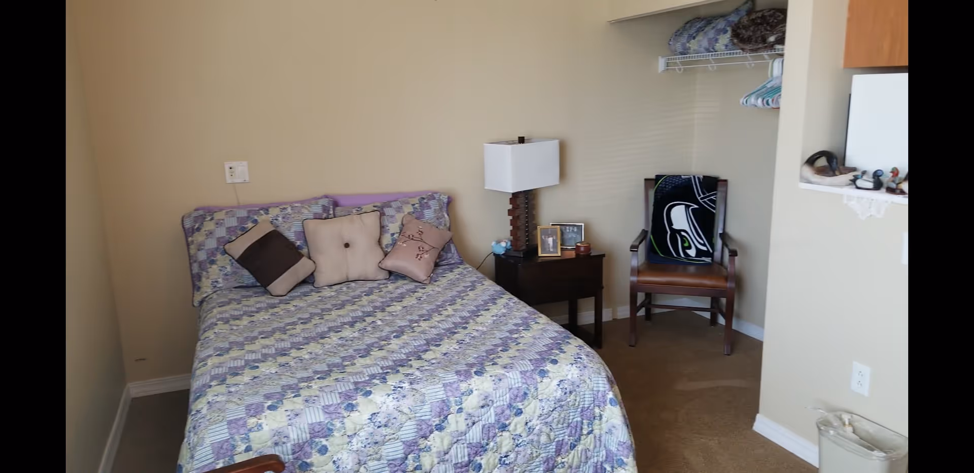 A small bedroom with a bed covered in a purple and white patterned quilt and several pillows. Next to the bed is a wooden nightstand with a lamp, two framed photos, and small decorative items. A wooden chair with a black and white blanket draped over the back is positioned near the corner. There is an open closet with a shelf holding folded blankets and hangers. The walls are beige, and the floor is carpeted.
