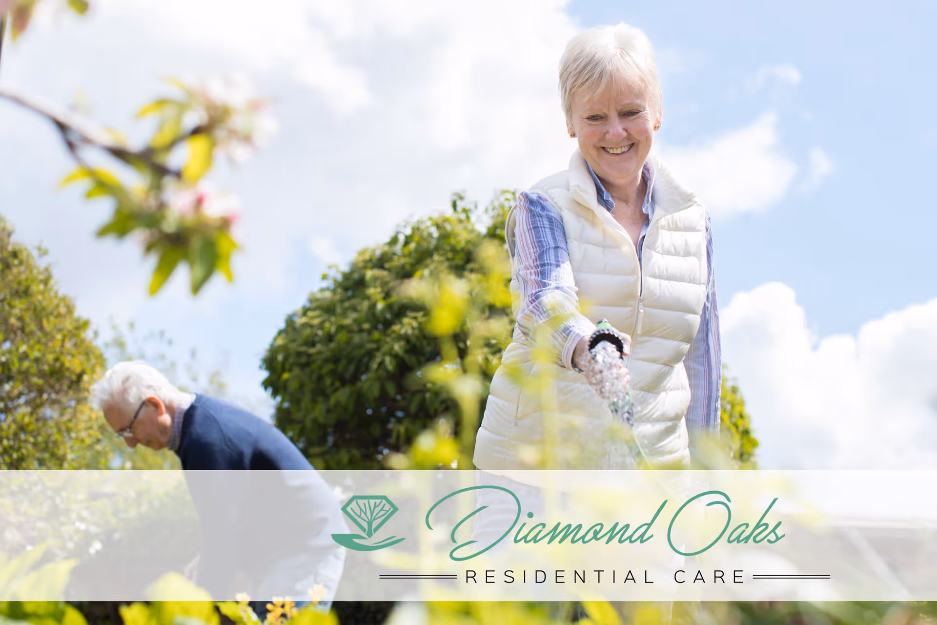 An elderly woman smiling and watering plants in a garden while an elderly man tends to plants in the background under a partly cloudy sky.