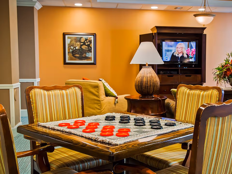 A cozy living room area with a wooden table set for a game of checkers with red and black pieces. Four striped upholstered chairs surround the table. In the background, there is a beige couch with colorful pillows, a wooden side table with a large lamp, a TV cabinet with a television showing a woman, and a framed abstract painting on an orange wall.