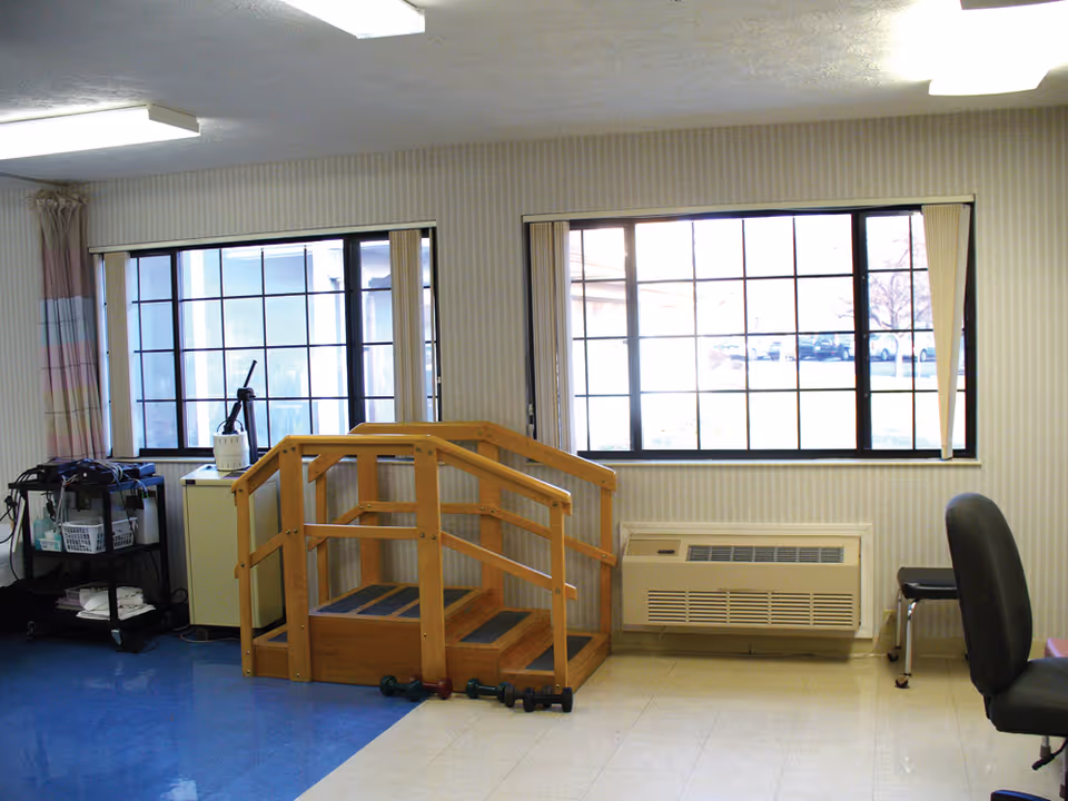 A room with two large windows, a small wooden staircase with handrails, a black office chair, a cart with medical or therapy supplies, and some small dumbbells on the floor.