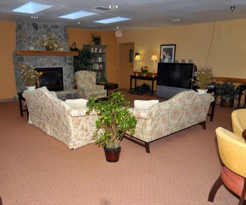 Cozy communal lounge with floral-upholstered sofas arranged around a coffee table, a stone fireplace, TV, and potted plants.
