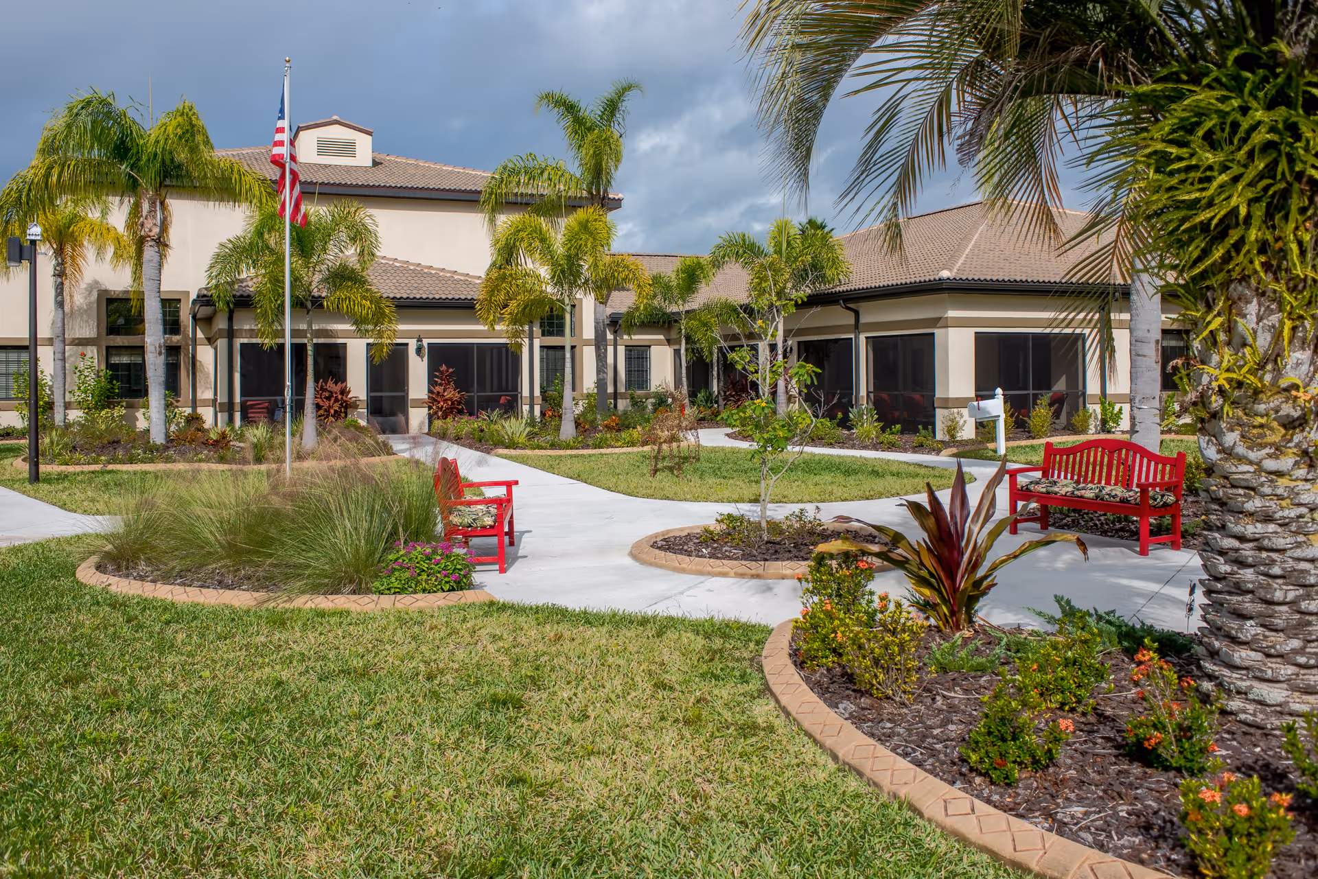 Outdoor view of Inspired Living Sun City Center showing a landscaped garden with palm trees, green grass, flower beds, and red benches along a paved walkway leading to a beige building with a tiled roof. An American flag is also visible on a flagpole near the center of the image.