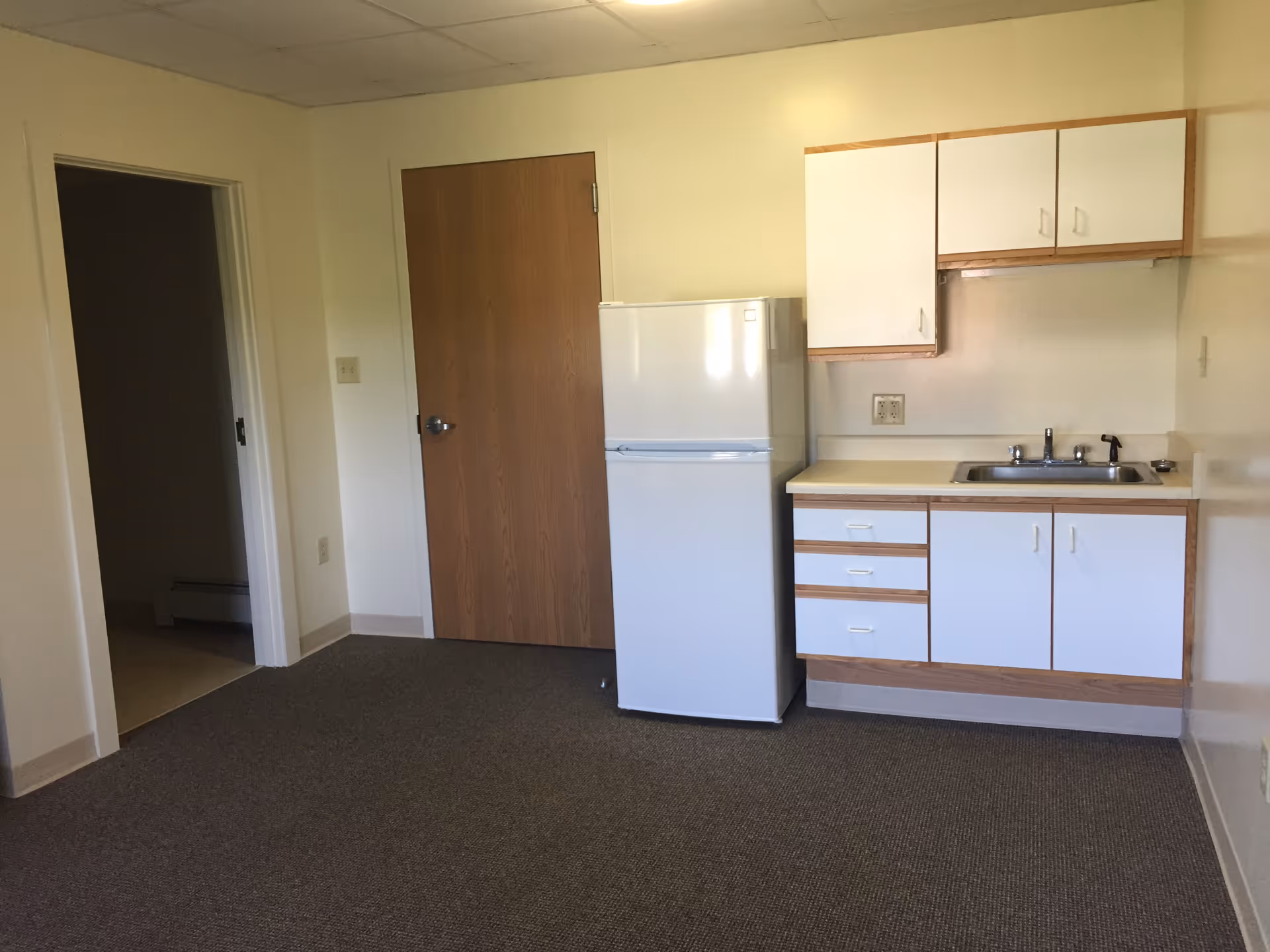 Small kitchenette area with a white refrigerator, white cabinets with wood trim, a sink, and a countertop. There is a closed wooden door and an open doorway leading to another room. The floor is carpeted and the walls are painted light yellow.