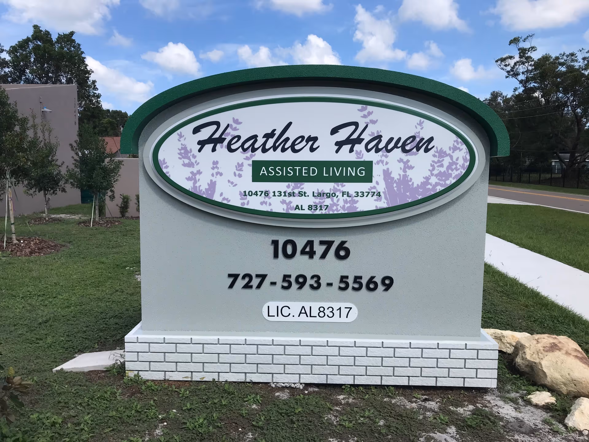 Outdoor sign for Heather Haven Assisted Living located at 10476 131st St. Largo, FL 33774 with contact number 727-593-5569 and license number AL 8317, surrounded by grass and trees with a road visible in the background under a partly cloudy sky.