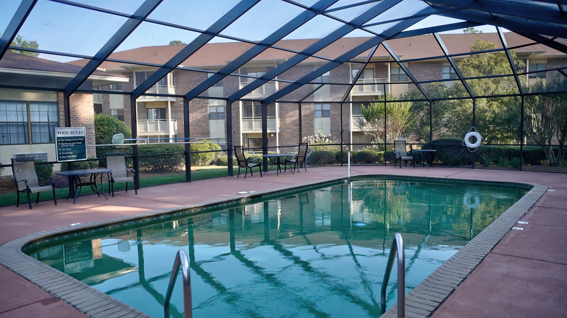Screened outdoor swimming pool with metal handrails, patio tables and chairs, and an apartment building visible behind the enclosure.