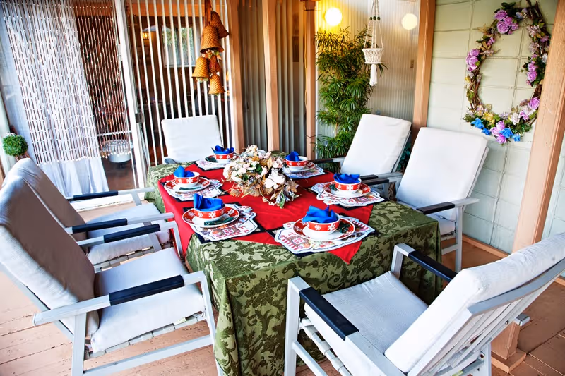 Outdoor dining area with a table covered by a green and red tablecloth, set with six place settings including plates, bowls, and blue napkins. The table is surrounded by six cushioned chairs. The area is decorated with a floral wreath on the wall, hanging bells, and potted plants, with a wooden slat wall and glass door in the background.