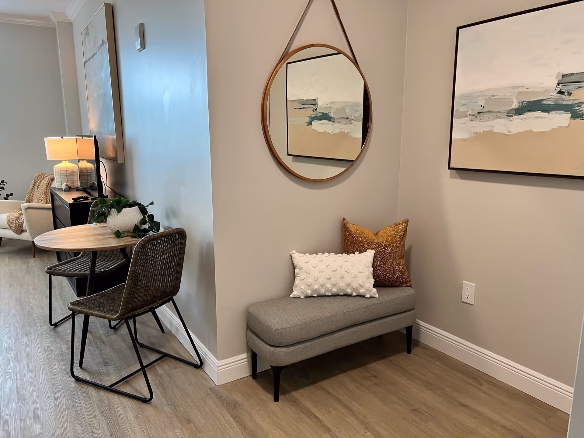 A cozy corner of a living area featuring a small gray upholstered bench with two decorative pillows, a round wall mirror hanging above it, and an abstract painting on the wall. To the left, there is a small round wooden table with a woven chair, a lamp on a black cabinet, and a beige armchair with a throw blanket in the background. The room has light wood flooring and neutral-colored walls.