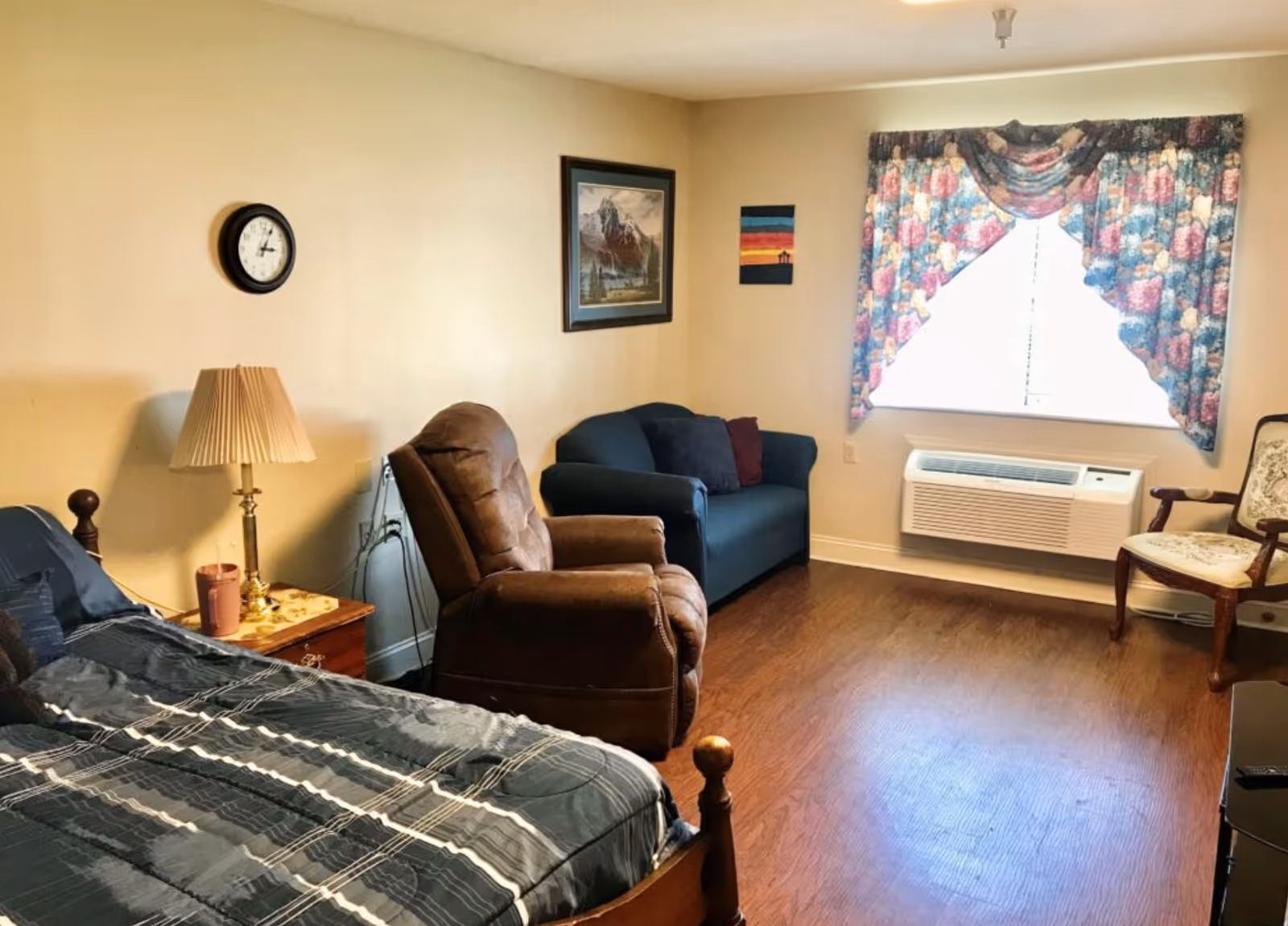 Cozy bedroom with a bed, recliner and small sofa facing a window with floral curtains and a wall-mounted air conditioner.