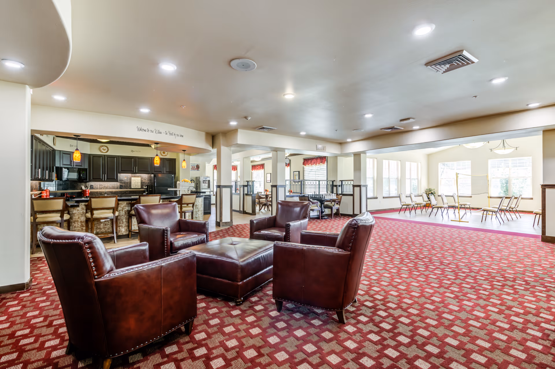 Spacious senior living common area with four leather armchairs around an ottoman, a bar/kitchen area in the background, and tables near large windows.