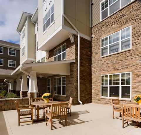 Outdoor patio area at The Greens at Hammonds Lane featuring wooden tables and chairs with yellow flower centerpieces, adjacent to a multi-story building with stone and siding exterior walls and multiple windows.