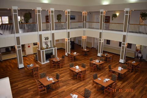 Spacious dining area with multiple wooden tables and chairs arranged neatly on a polished wooden floor. The room features a two-story design with white railings and columns, potted plants hanging from the upper level, and soft lighting. A fireplace is visible on one side of the room.