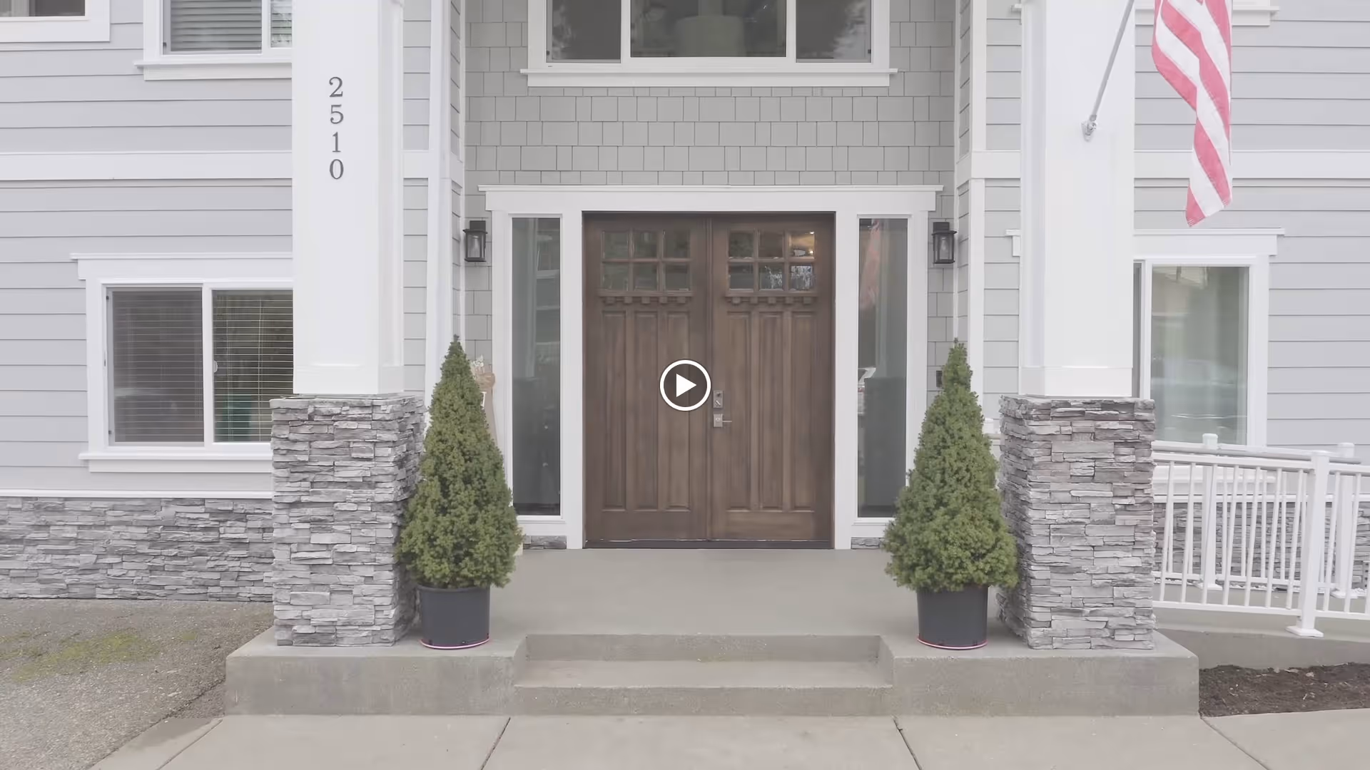 Front entrance of a senior living facility with double wooden doors, flanked by two potted evergreen shrubs. The building exterior features gray siding with stone accents on the lower walls and white trim. The address number 2510 is displayed vertically on a white column to the left of the door, and an American flag is visible on the right side.