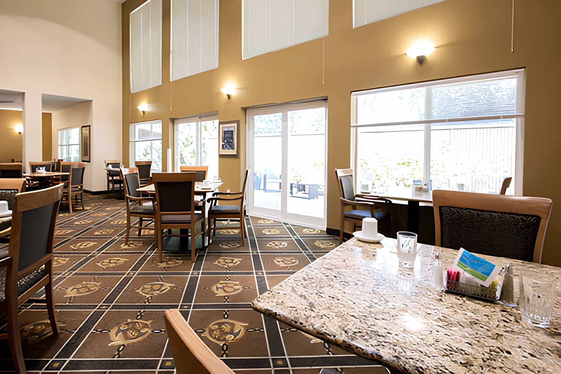 A bright dining room in a senior living facility with multiple wooden tables and chairs arranged on a patterned carpet. Large windows and sliding glass doors allow natural light to fill the space. The walls are painted beige with wall-mounted lights, and the tables are set with glasses and napkins.