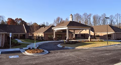 Exterior view of a single-story senior living facility with a circular driveway and a covered entrance. The building has a brick facade and a small cupola on the roof. Leafless trees are visible in the background under a clear sky.