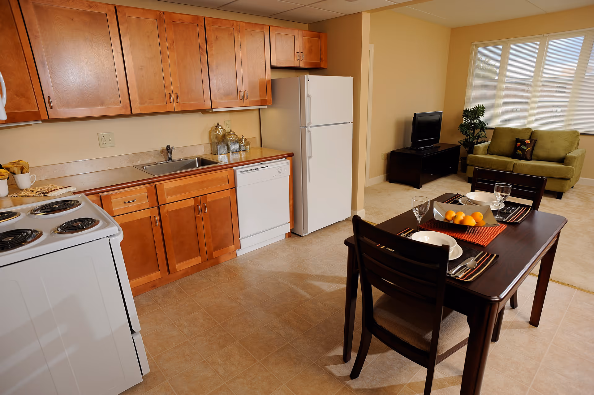 A bright and clean kitchen and dining area in a senior living facility. The kitchen features wooden cabinets, a white refrigerator, dishwasher, stove, and a sink. Adjacent to the kitchen is a dark wooden dining table set for two with plates, glasses, utensils, and a bowl of oranges. In the background, there is a living area with a green sofa, a small TV on a stand, a plant, and large windows letting in natural light.