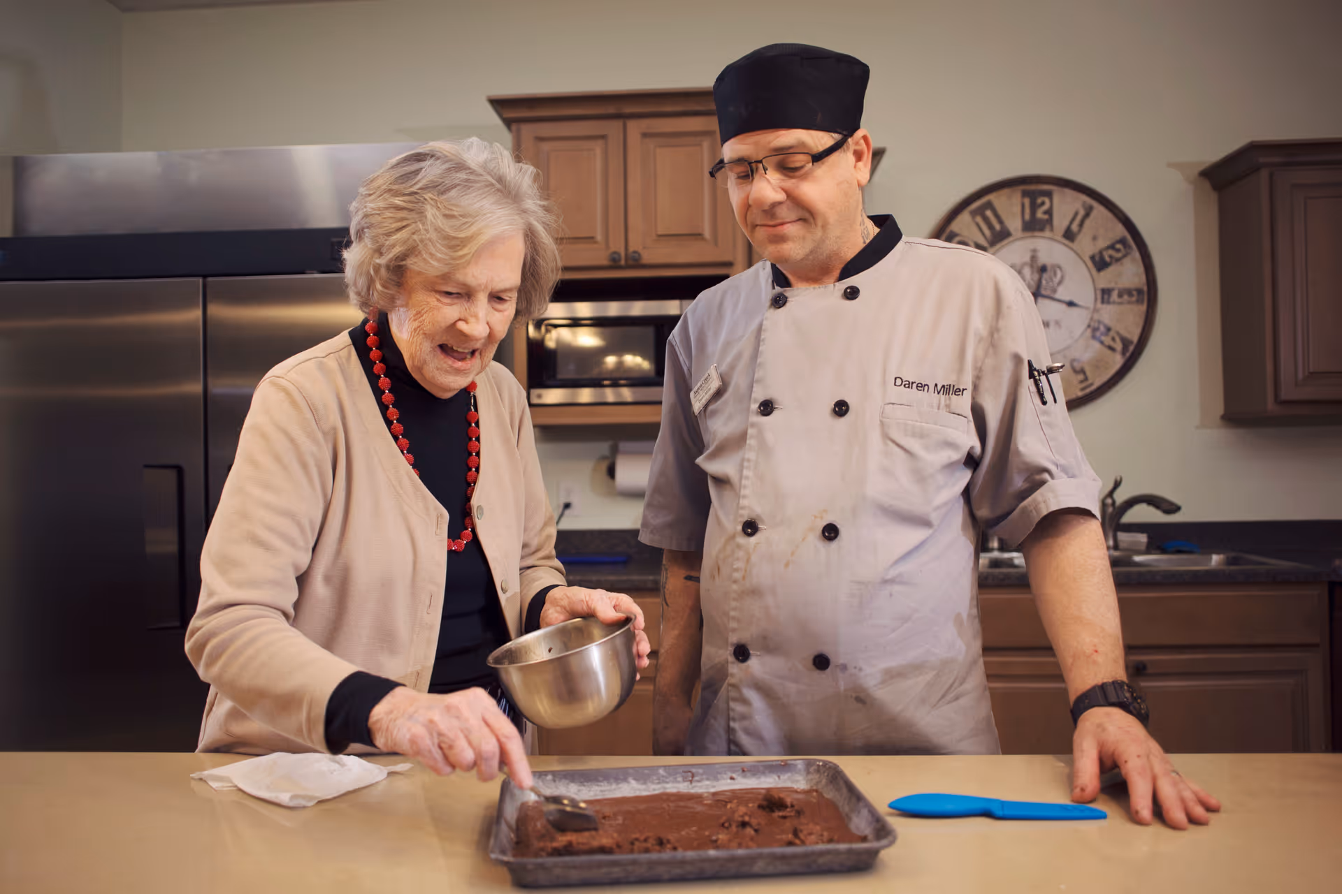An elderly woman and a chef in a kitchen preparing a chocolate dessert together. The woman is holding a small metal bowl and using a spoon to spread the dessert in a baking tray, while the chef watches and smiles. The kitchen has wooden cabinets, a large refrigerator, and a wall clock in the background.