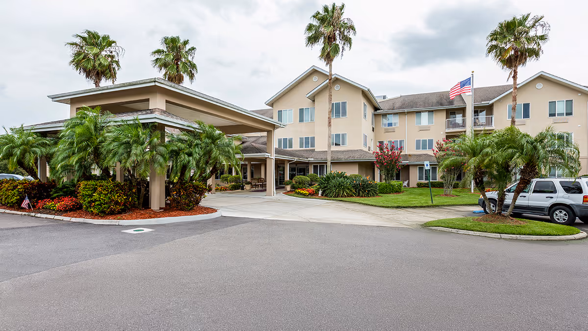 Front entrance of a beige three-story senior living building with a covered porte-cochère, palm trees, landscaping, and parked cars.