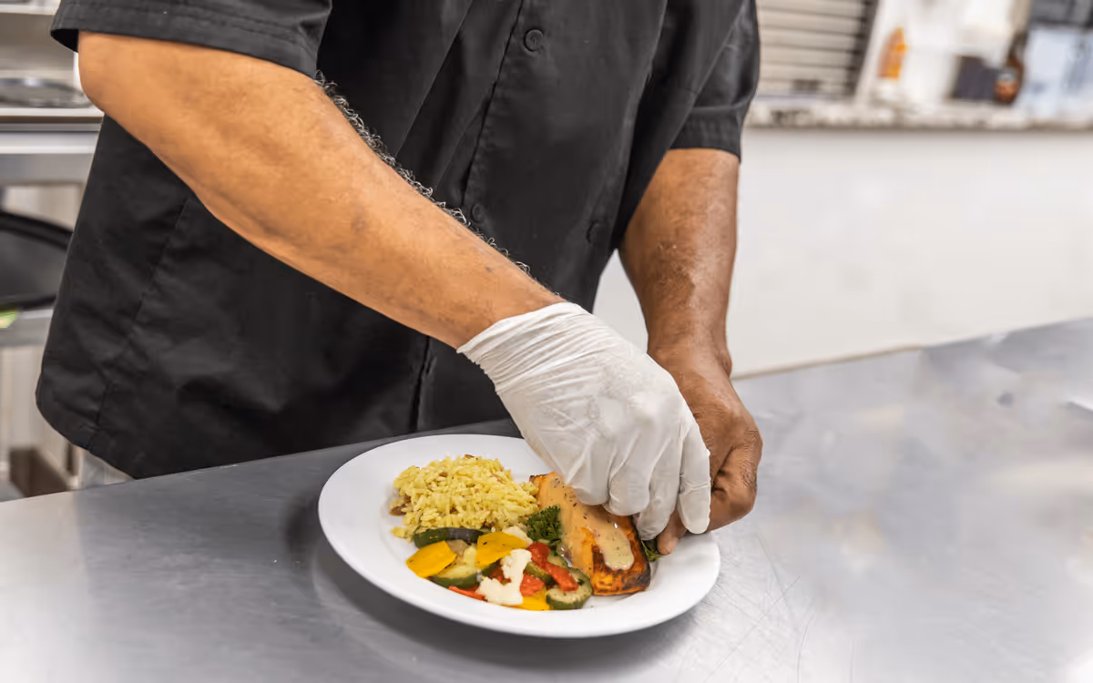 A person wearing a black chef's coat and white gloves is plating a meal consisting of grilled salmon with sauce, yellow rice, and mixed vegetables on a white plate in a kitchen setting.