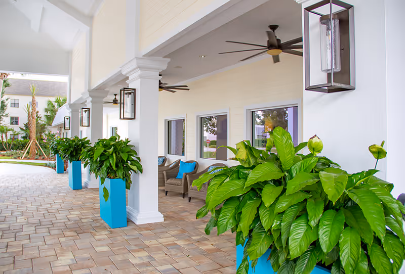 Covered outdoor walkway at Buena Vida Estates with large green plants in blue planters along white columns, ceiling fans, and wicker chairs with blue cushions near windows.