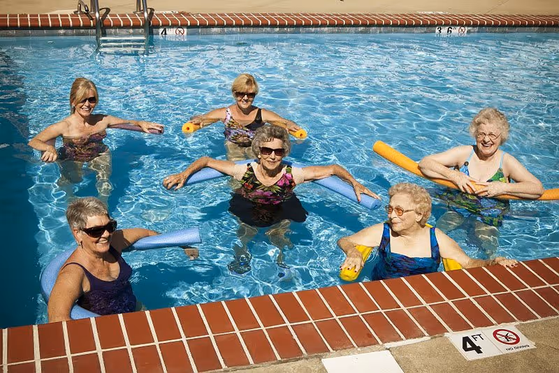 Six elderly women in swimsuits enjoying a water exercise session in an outdoor swimming pool, each using a colorful pool noodle for support. The pool has a brick edge and depth markers visible.
