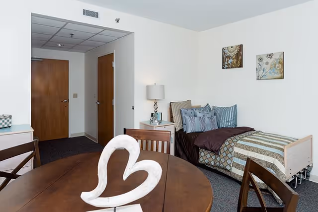 A cozy senior living bedroom with a single bed made with patterned bedding and multiple pillows. Next to the bed is a nightstand with a lamp and decorative items. There is a wooden dining table with chairs in the foreground, featuring a white heart-shaped sculpture on it. The room has white walls with two framed artworks hanging above the bed and two wooden doors visible in the background.