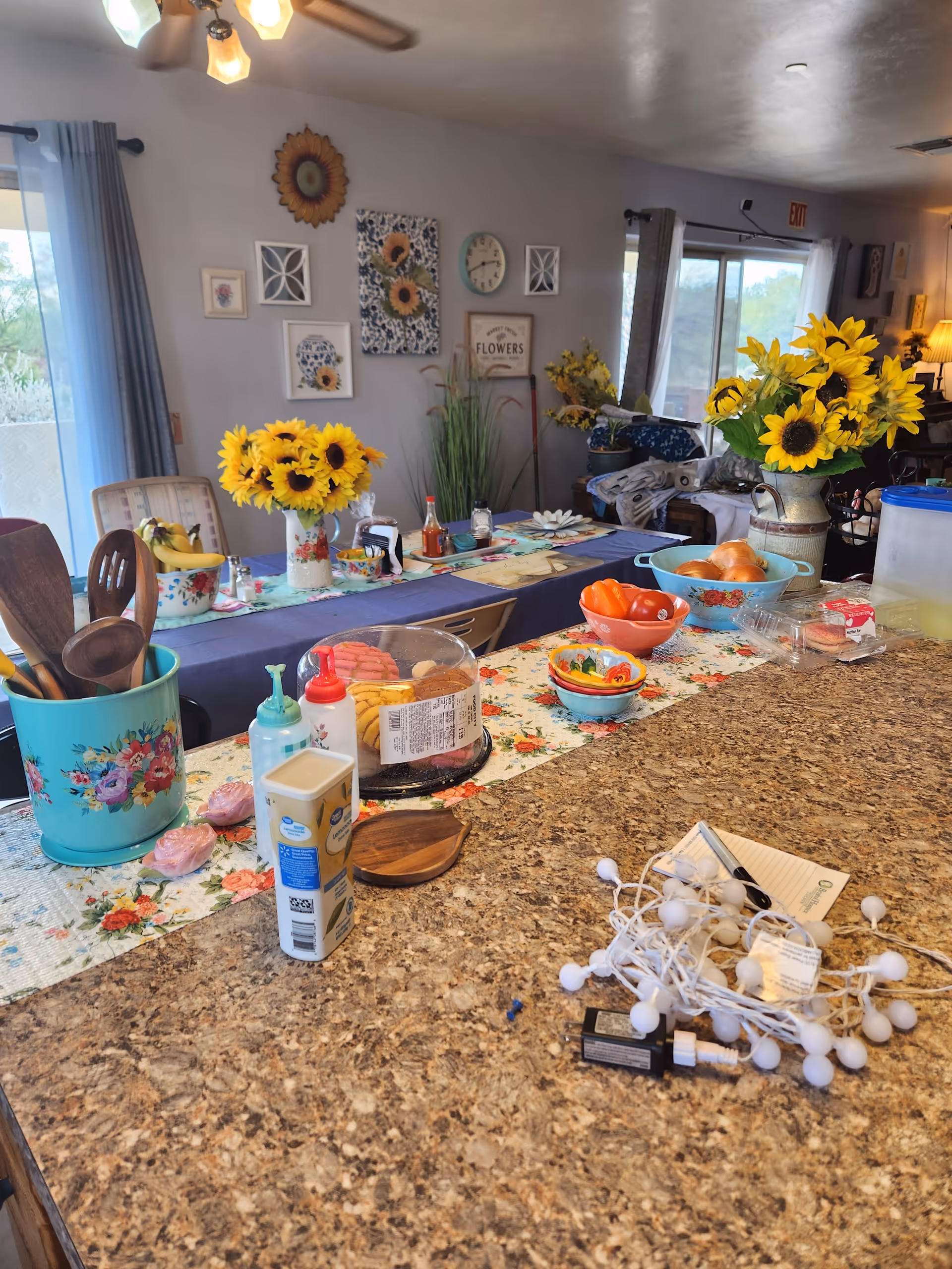 A cozy interior space with a granite countertop in the foreground holding various items including kitchen utensils in a floral container, condiments, a container of cookies, a bowl of tomatoes, and a string of white lights. Behind the counter is a long dining table decorated with a floral runner, sunflower bouquets, and various condiments. The walls are adorned with sunflower-themed artwork and a clock. Large windows with curtains allow natural light into the room.