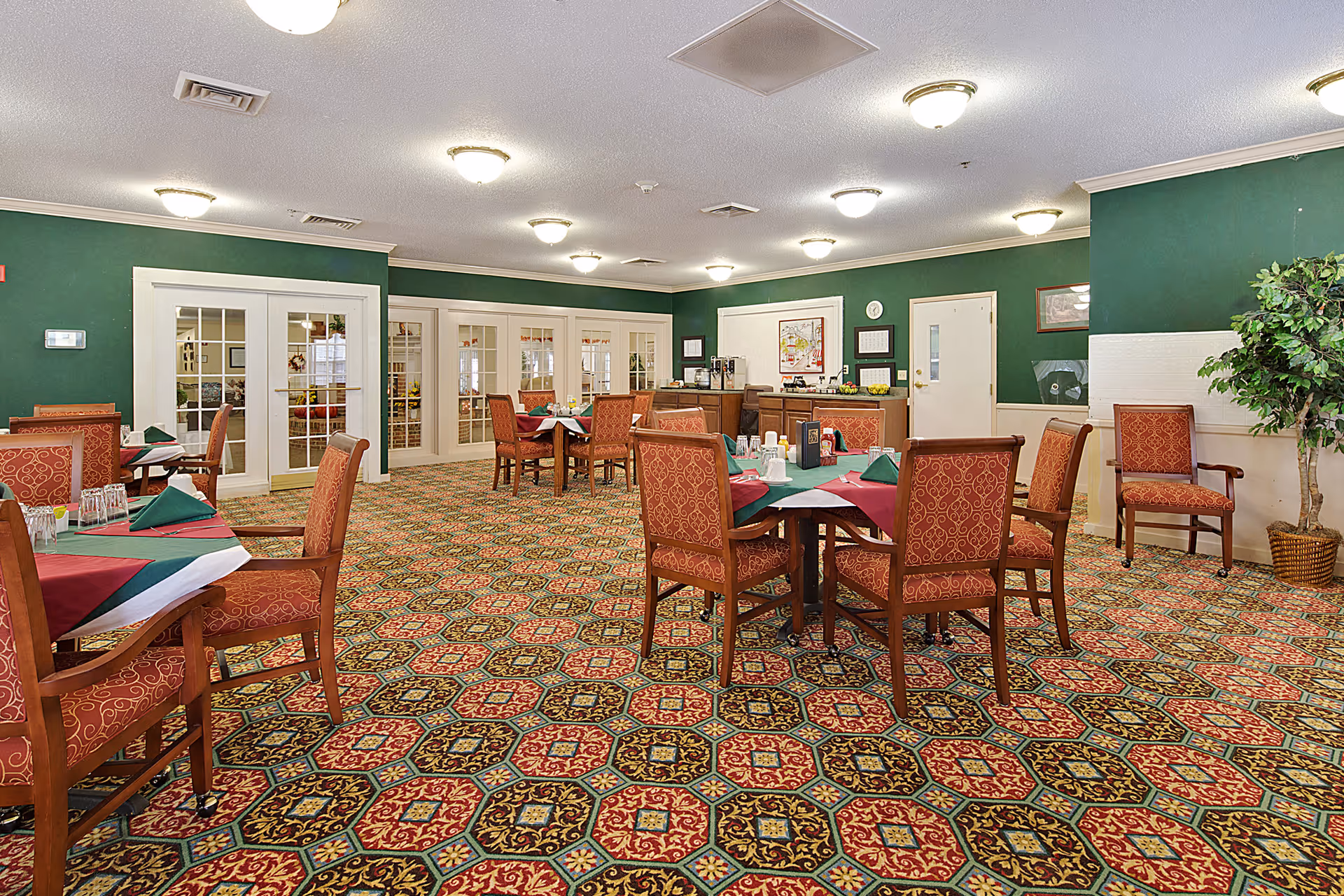 Dining room with patterned carpet, multiple tables set with napkins and upholstered chairs against green walls and French doors.