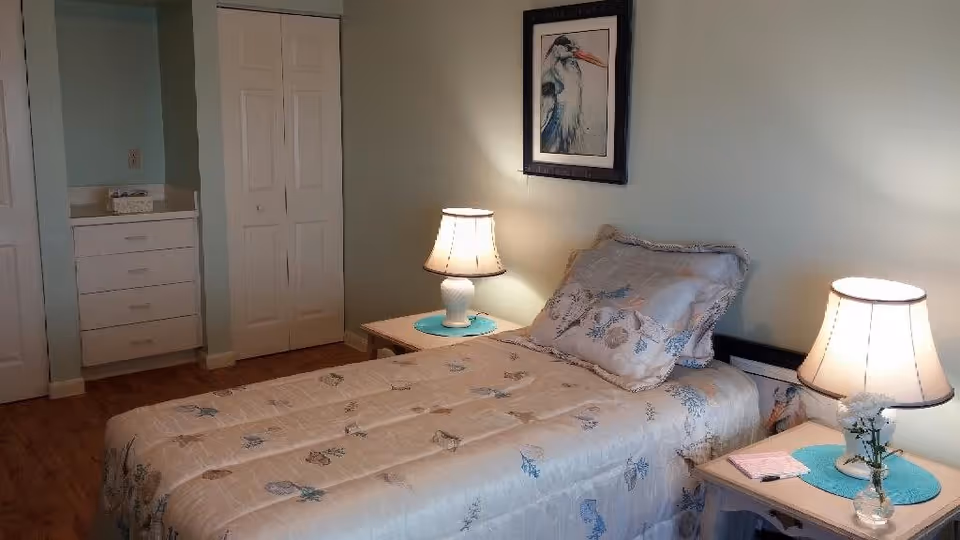 A cozy bedroom with a single bed covered in a light-colored floral comforter and matching pillow. There are two bedside tables, each with a lamp and a blue placemat. A framed picture of a bird hangs on the wall above the bed. The room has light-colored walls and wooden flooring. In the background, there is a white closet with double doors and a small built-in dresser with drawers.