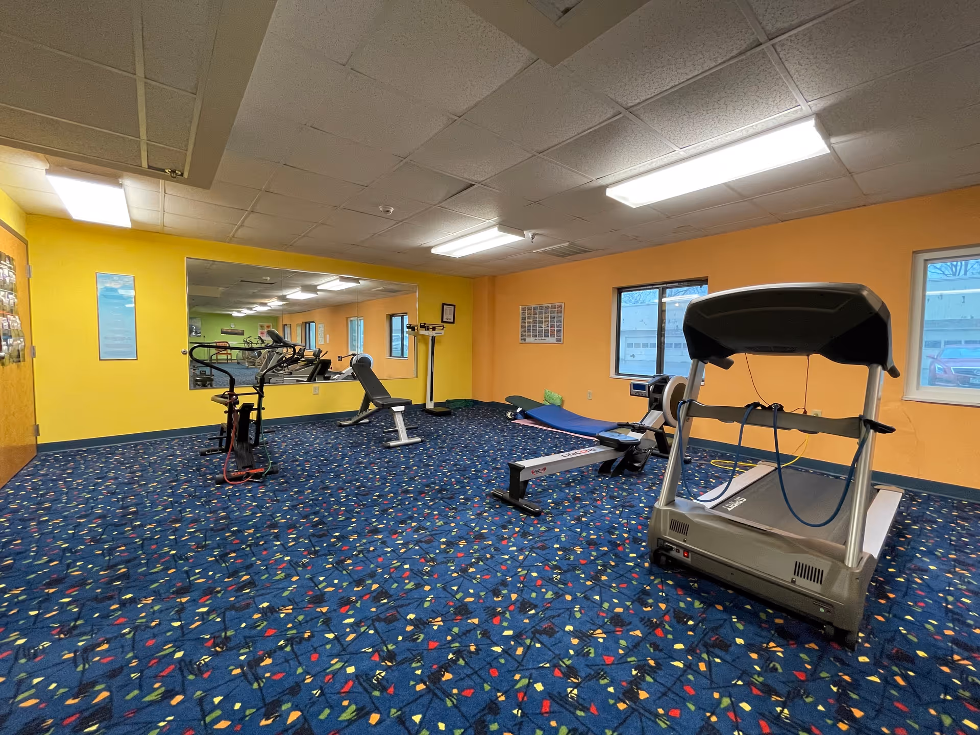 A fitness room with exercise equipment including a treadmill, rowing machine, stationary bike, and weight bench. The room has bright yellow and orange walls, a large mirror on one wall, blue carpet with colorful patterns, and two windows showing an outside view.