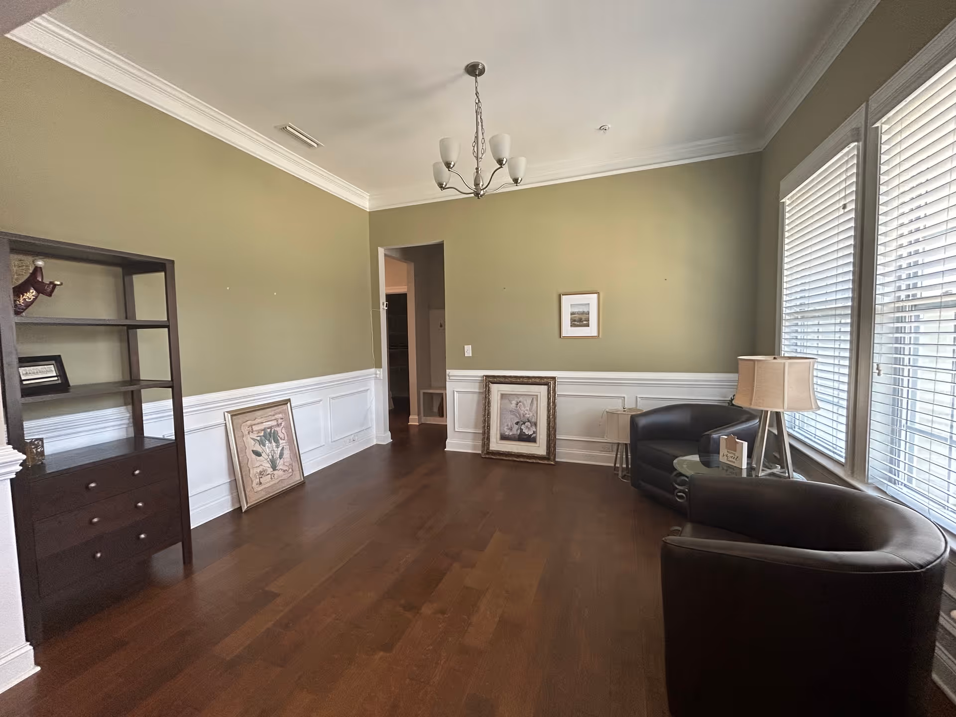 A small sitting area with two black leather armchairs, a round glass-top side table with a lamp and a small sign, and two framed pictures leaning against a green wall with white wainscoting. There is a dark wooden shelving unit on the left side and large windows with white blinds on the right, letting in natural light. The floor is wooden and a chandelier hangs from the ceiling.