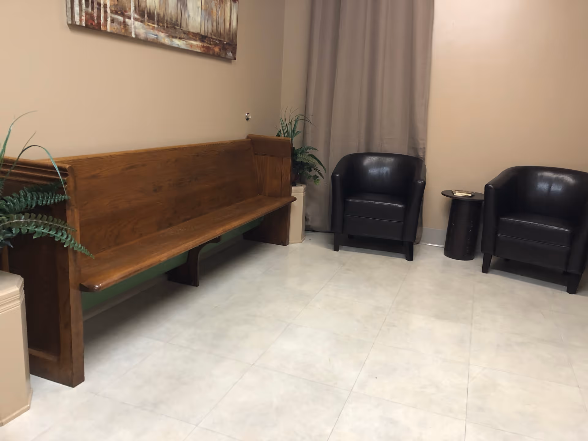 A waiting area with a long wooden bench on the left, two black leather armchairs on the right, a small round wooden side table between the chairs with a book on it, beige walls, a large curtain covering a window, and a painting hanging above the bench. There are also two potted plants near the bench and armchairs.