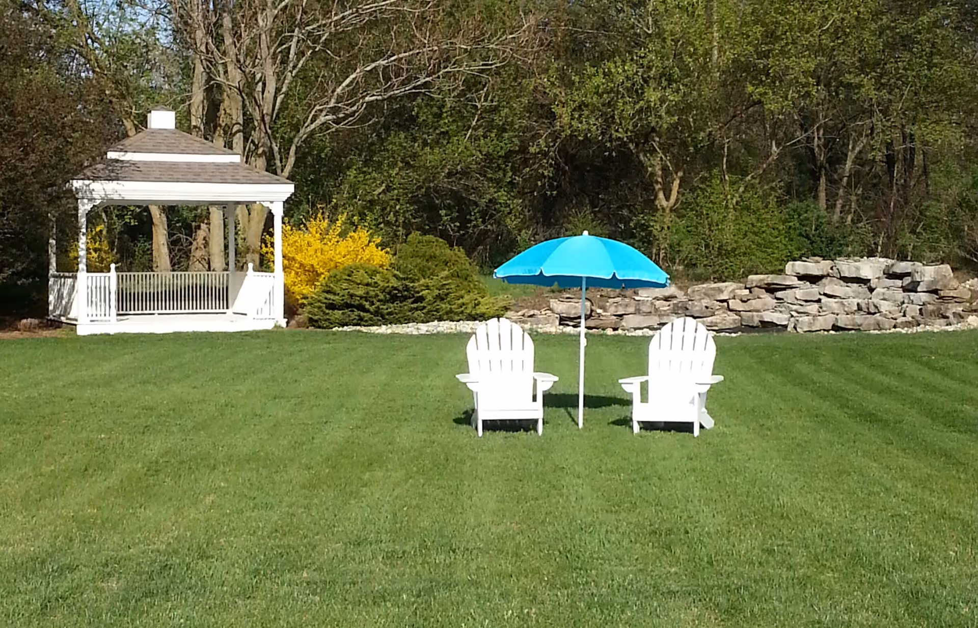 A grassy outdoor area with two white Adirondack chairs facing away, positioned under a blue umbrella. To the left, there is a white gazebo surrounded by green bushes and trees in the background.