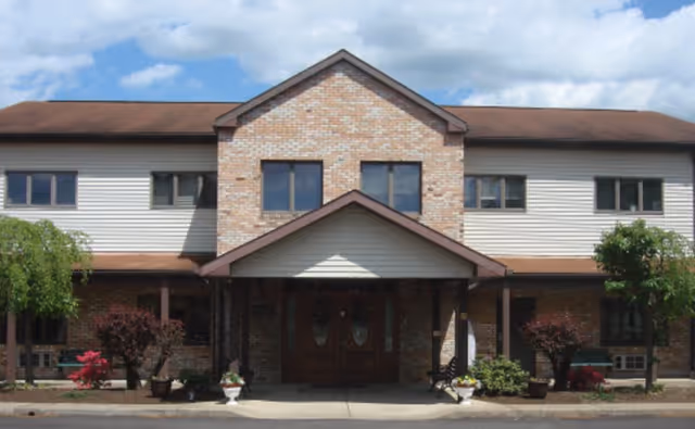 Front exterior view of a two-story building with a brick and siding facade, a peaked roof, and a covered entrance with double wooden doors. There are small trees and shrubs planted along the front of the building under a partly cloudy sky.