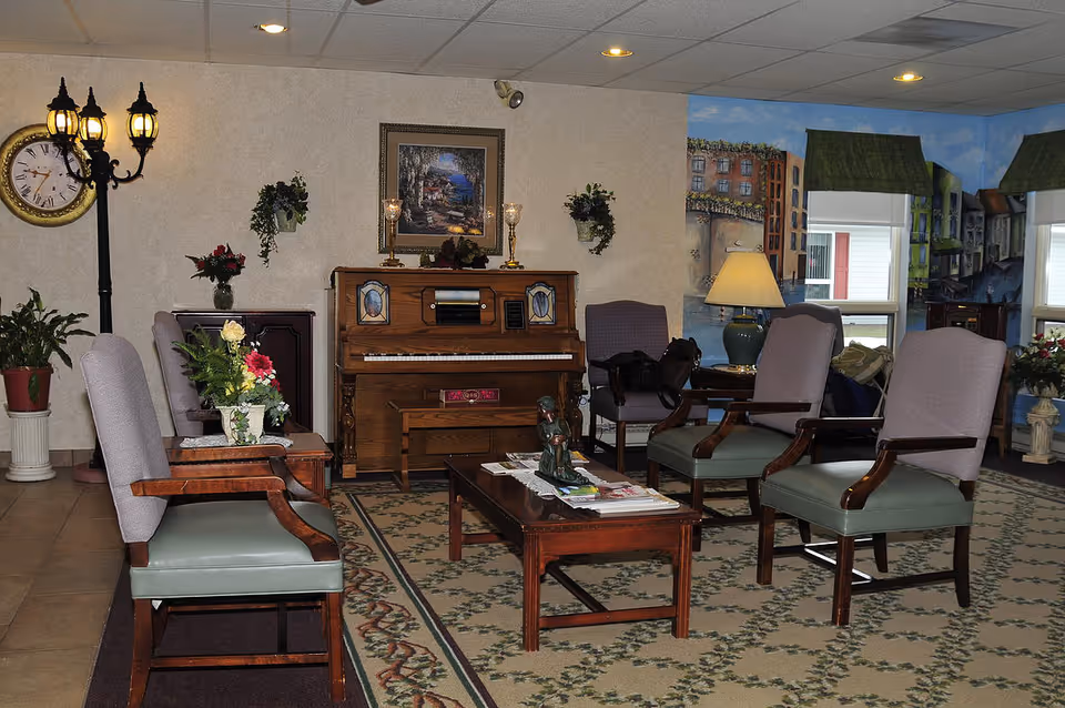 A cozy living room area with several wooden chairs upholstered in green and purple fabric arranged around a wooden coffee table with magazines and a small statue. A wooden piano with decorative panels is against the wall, flanked by two wall-mounted plants and a framed painting above it. A floor lamp with three lantern-style lights and a large clock are on the left side. The walls feature a mural of a canal scene with buildings and greenery, and there are windows with green valances letting in natural light.