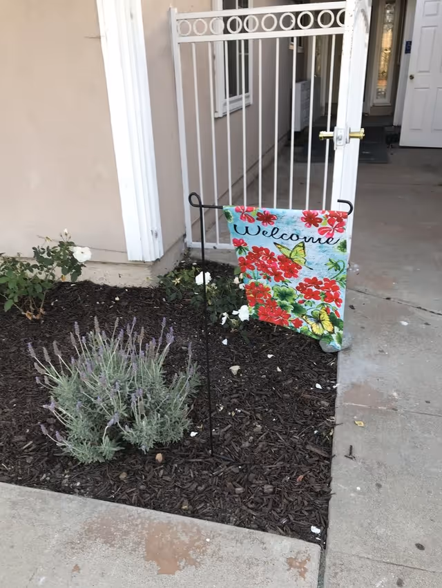 A small garden bed with mulch, a lavender plant, and white flowers next to a beige building wall. A decorative garden flag with red flowers, green leaves, yellow butterflies, and the word 'Welcome' is displayed on a black metal stand. Behind the garden bed is a white metal gate and a concrete walkway leading to a white door.