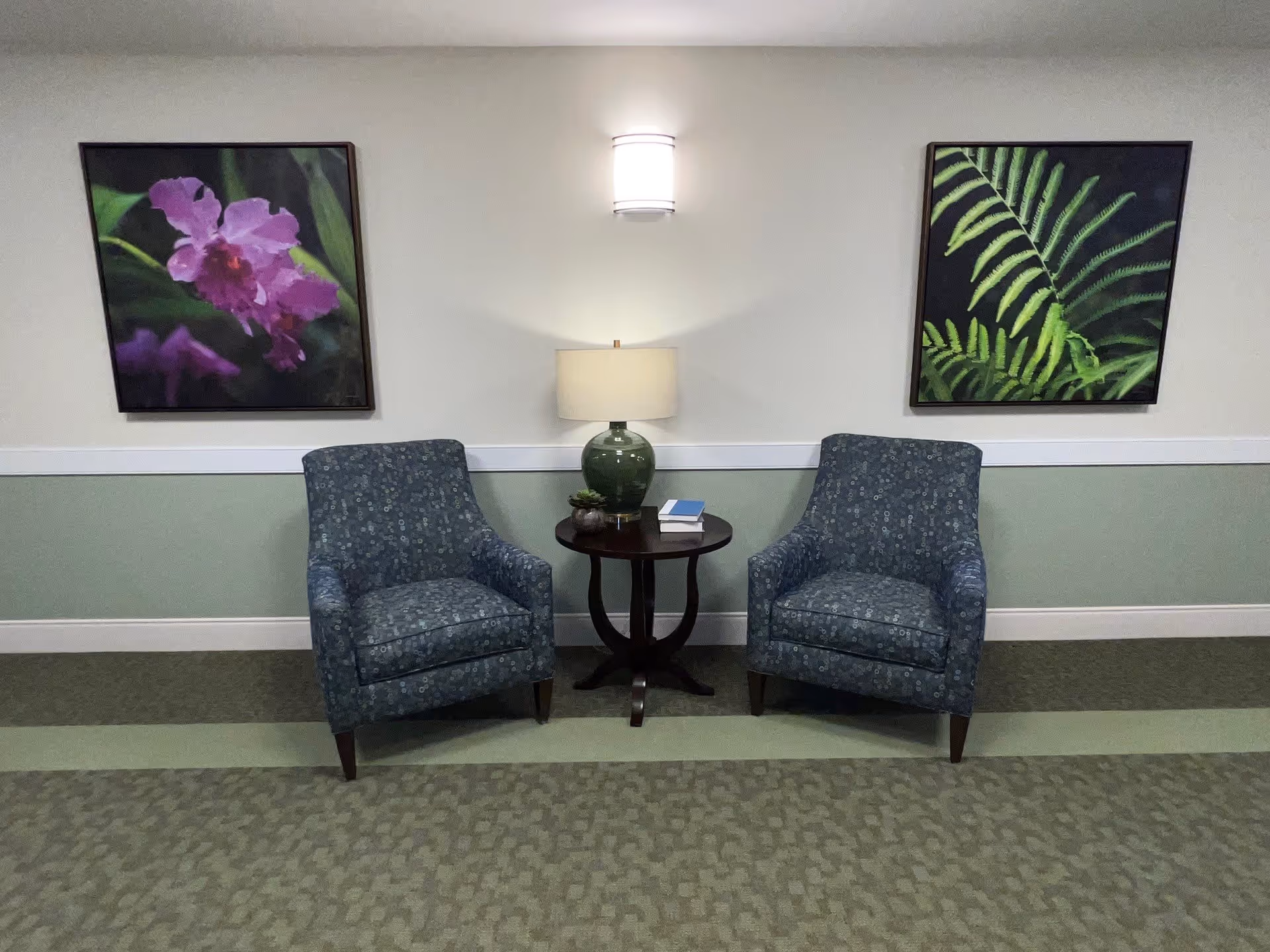 Two blue patterned armchairs with wooden legs are positioned on either side of a small round wooden table. On the table is a green glass lamp with a beige lampshade, two stacked books, and a small potted plant. The wall behind the chairs is painted light green on the lower half and white on the upper half, with a white chair rail separating the colors. Above the lamp is a wall-mounted light fixture. On the wall, there are two framed paintings: one of purple flowers on the left and one of green fern leaves on the right.