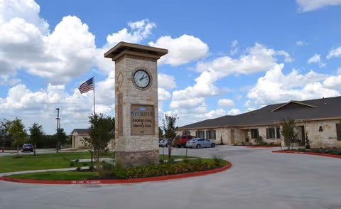 Exterior view of New Haven Assisted Living and Memory Care of Kyle facility with a stone clock tower sign displaying the facility name, an American flag, parked cars, and a partly cloudy sky.