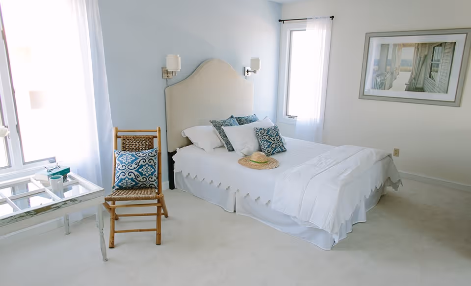 A bright and airy bedroom with a neatly made bed featuring white linens and decorative blue and white pillows. A straw hat rests on the bed. Next to the bed is a wooden chair with a blue patterned cushion. A glass-top table with a cup and a book is positioned near a window with sheer white curtains. A framed picture hangs on the wall above the bed.