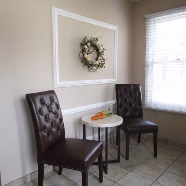 A small seating area with two dark brown tufted leather chairs and a round marble-top table between them. On the table, there are three carrots with green tops. The wall behind the chairs is beige with white trim and a decorative wreath hanging in a framed section. A window with white blinds is on the right side, letting in natural light.