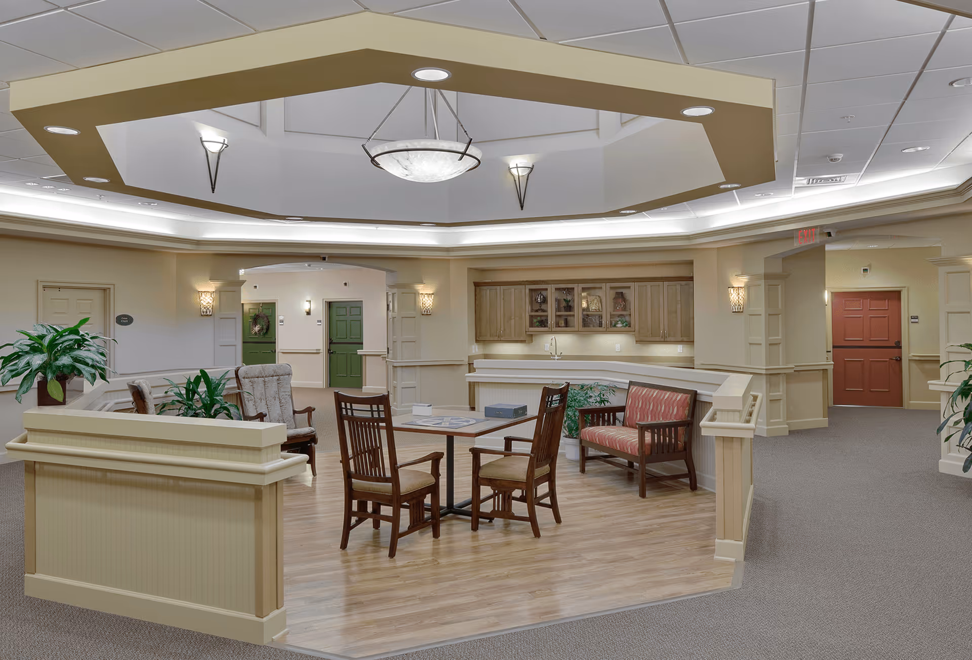 A well-lit common area in a retirement community featuring a small seating arrangement with wooden chairs and a cushioned bench around a square table. The space has a decorative ceiling with recessed lighting and a central hanging light fixture. There are plants placed around the area, and wooden cabinets with glass doors are mounted on the back wall. Several doors and hallways lead to other parts of the facility.