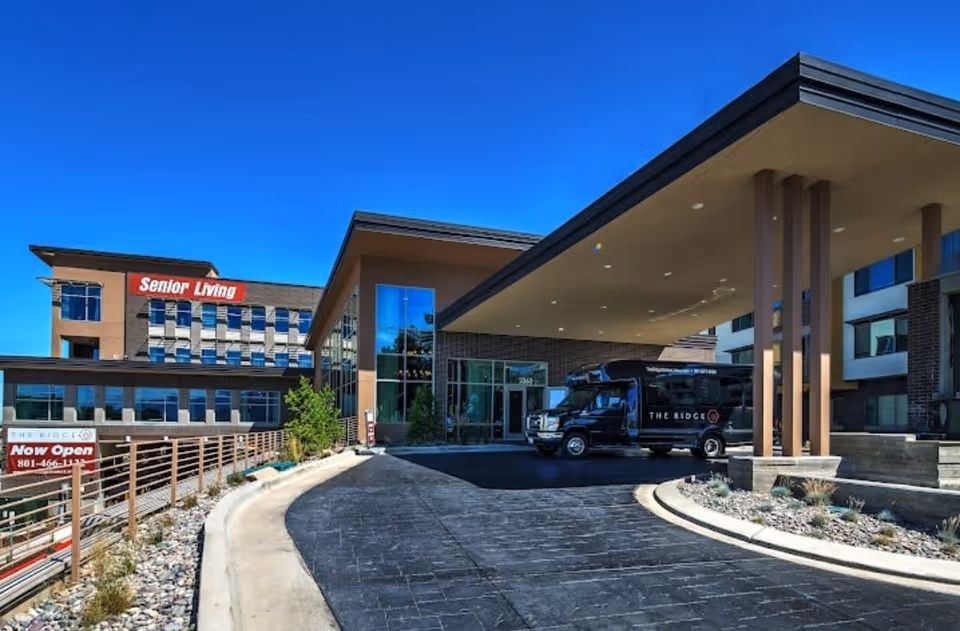 Exterior view of The Ridge Foothill senior living facility with a modern building, a covered entrance with pillars, a black shuttle van parked under the canopy, and clear blue sky. A sign near the entrance reads 'Now Open' with a phone number.