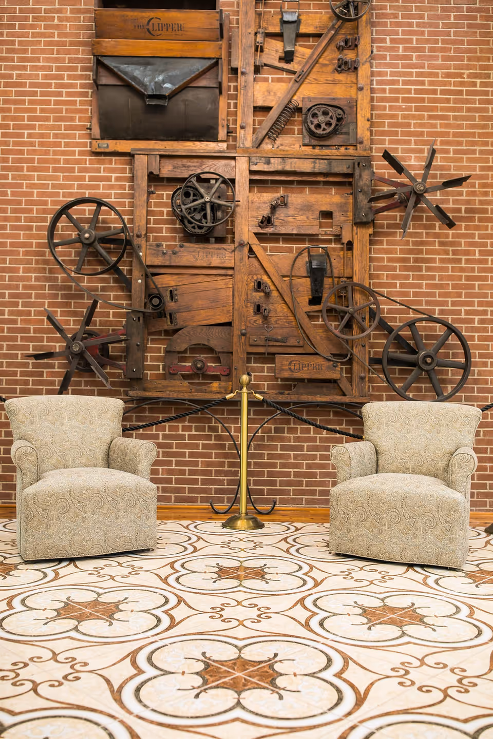 Two patterned armchairs placed on a decorative tiled floor in front of a brick wall featuring a large wooden mechanical wall art installation with gears and wheels.